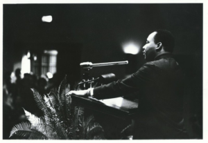 A B&W photo of Dr. Martin Luther King, Jr at a podium with a bright light shining just to the left of his face and a fern to the left of the podium.